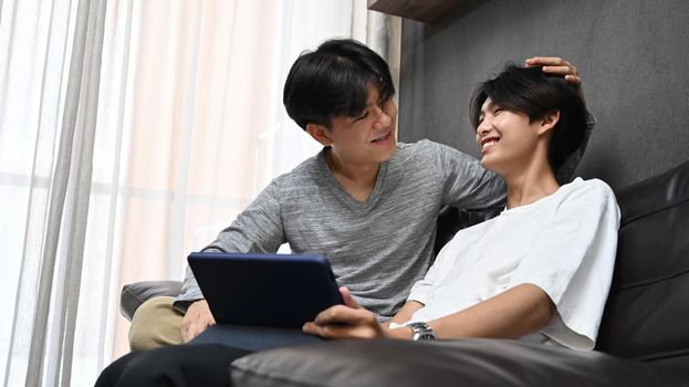 Loving young gay couple using digital tablet together in living room. Concept of sexual freedom and equal rights for LGBT community.