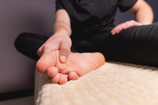 A man sitting on a massage table does self-massage of his feet with his own hands.