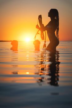 Silhouette frame. A young slim sexy female kitesurfer with a plank and a kiteboard stands in the water in the shallow water at sunset. Water sports. Stylized frame.