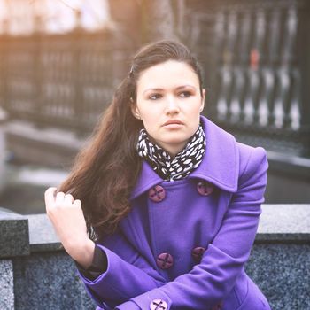 Beautiful girl sitting on a bench in fialetovom coat in clear time of the day, wondering and looking in the direction of hair straightening