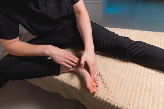 A man sitting on a massage table does self-massage of his feet with his own hands.