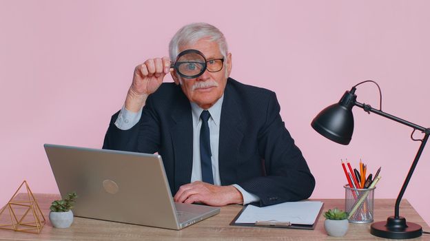 Investigator researcher scientist senior businessman holding magnifying glass near face, looking into camera with big zoomed funny eye, searching, analysing sits alone at workplace desk at pink office