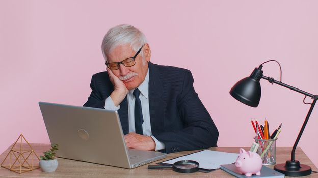 Tired sleepy senior business man falling asleep sit at office desk with computer, overworked lazy bored grandpa worker feel fatigue close eyes sleeping on hand exhausted of work deprived concept
