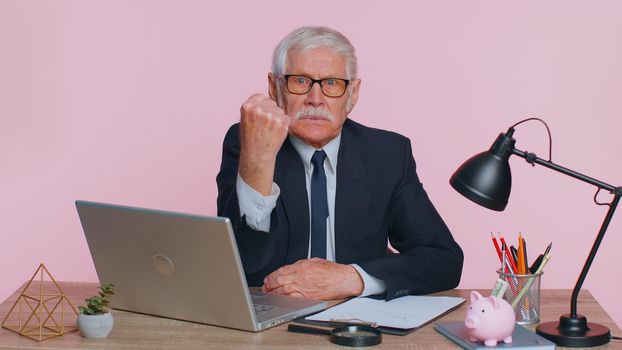 Aggressive angry senior businessman trying to fight at camera, shaking fist, boxing with expression, punishment sits at workplace desk at office. Elderly man isolated indoor on pink studio background