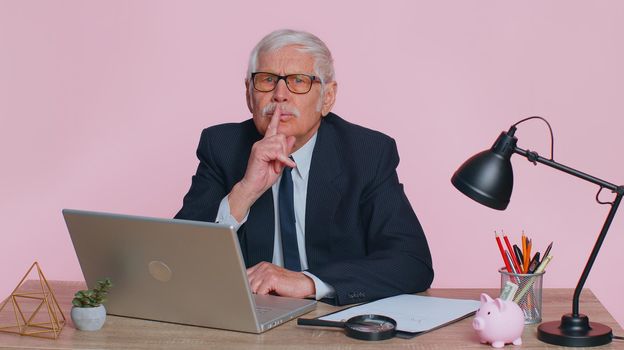 Shh be quiet please. Senior businessman in suit presses index finger to lips makes silence gesture sign do not tells secret sits at workplace desk at office. Elderly man on pink studio wall background