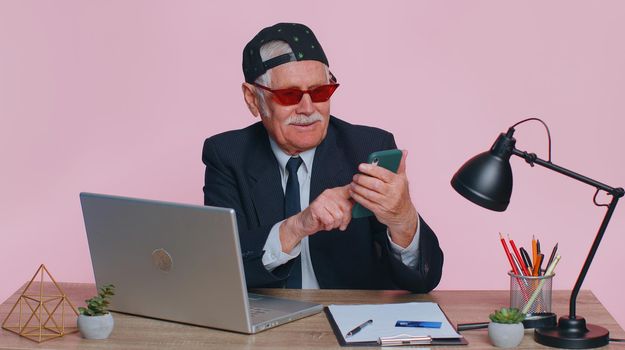 Funny stylish businessman using smartphone typing news post on web sms message, browsing, searching, addiction of social networks at office. Senior grandfather man studio isolated on pink background