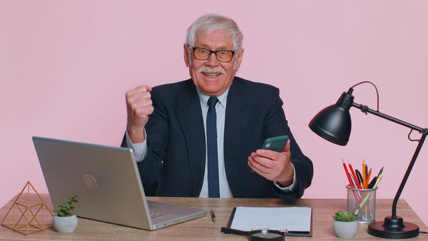 Happy excited joyful senior businessman use smartphone typing browsing say wow yes, great big win good news shocked by sudden victory at office. Elderly man isolated indoor on pink studio background