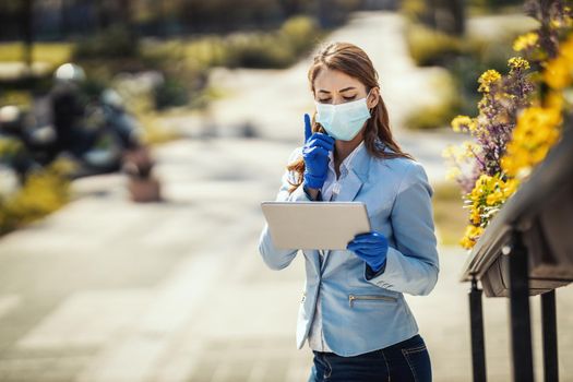 Young business woman with protective mask on her face is looking something on her digital tablet outdoors.