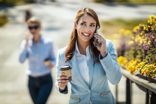 Young smiling businesswoman is walking in front of office and talking on the smartphone during coffee break.