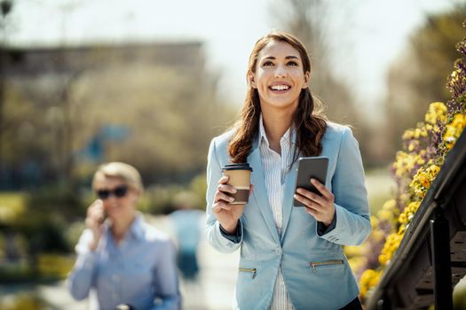 Young smiling businesswoman is standing in front of office and using smartphone during coffee break.