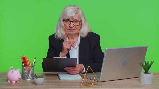 Senior businesswoman analysing paperwork documents shakes finger saying no, be careful, scolding and giving advice to avoid danger mistake disapproval sign at office. Elderly woman on green chroma key
