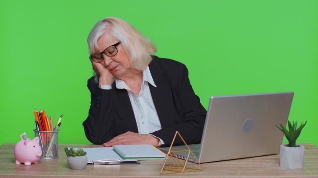 Tired sleepy senior business woman falling asleep sit at office desk with computer, overworked lazy bored grandpa worker feel fatigue close eyes sleeping on hand exhausted of work deprived concept