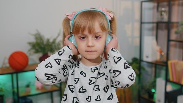 Close-up portrait of little happy child girl dancing on sofa while listening music in headphones in cozy living room. Small cute teen school kid at home alone dancing, singing and moving to the rythm
