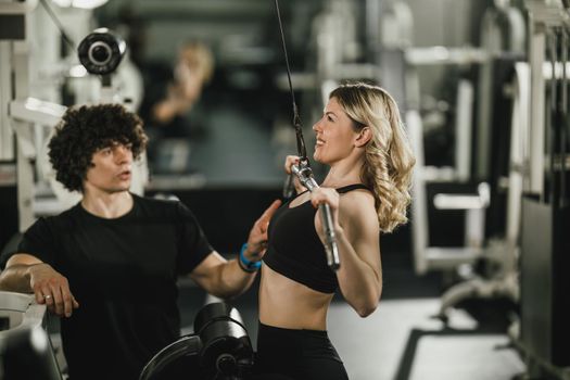 A young muscular woman is doing training at machine supporting by coach in the gym.