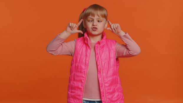 Funny joyful sincere little girl 6 years old in vest making playful silly facial expressions and grimacing, fooling around showing tongue. Young toddler child isolated alone on orange wall background