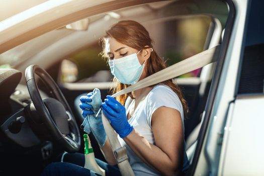 A young woman with a mask on her face and protective gloves on her nands wipes a car belt in her car with a cloth with disinfectant liquid.