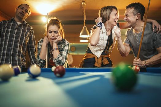 Young cheerful friends are playing billiards in bar after work. A couple that wins fame and the losers are in a bad mood.