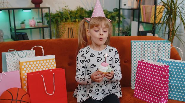 Young little children girl sitting on sofa with lots of gift presents, celebrating birthday party anniversary at home alone. Happy toddler child kid holding cake, making a wish and blowing one candle