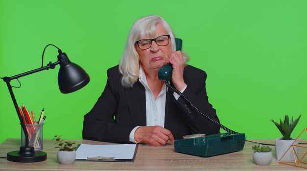 Crazy senior businesswoman talking on wired vintage telephone of 80s, fooling, making silly faces, humor comic, sits at workplace desk at office. Elderly woman isolated on green chroma key studio