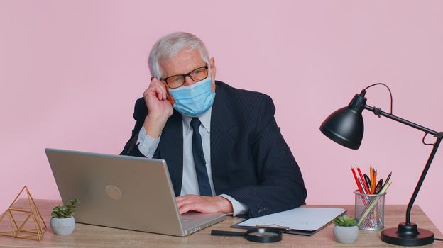 Sad unhappy senior businessman putting on face hygienic medical mask to prevent respiratory infection, airborne epidemic flu disease, contagious Coronavirus Covid-19 at workplace desk at pink office