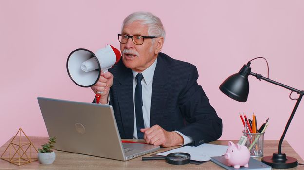 Cheerful senior old business man manager screaming in loudspeaker megaphone horn, discounts news and sales, Hurry up, announcement, advertising. Grandfather freelancer sitting at laptop in pink office