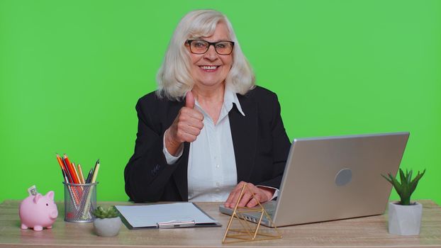 Senior old business woman employee using laptop computer notebook, raises thumbs up agrees with something or gives positive reply recommends advertisement likes good sits at green chroma key office