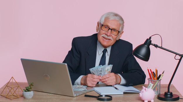 Senior business man accountant or banker making calculations at workplace desk. Elderly grandfather counting money dollar cash banknotes income earnings, working at office on pink studio background