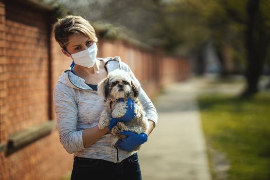 A woman in a medical protective mask is spending time with her dear cute little Shih Tzu dog on the city street path during flu virus outbreak and coronavirus epidemic.