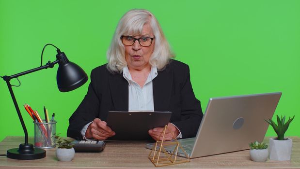 Oh my God Wow. Senior business woman, using laptop, analysing paperwork documents raising hands in surprise, shocked by sudden victory, good news. Elderly freelancer working at green chroma key office