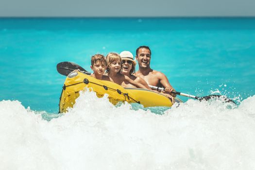 Happy family is enjoying a ride in rubber kayak and waves are splashing them at tropical ocean water during summer vacation.