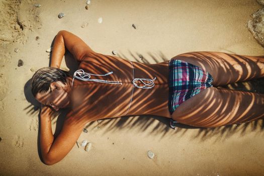 A beautiful young woman is relaxing on the beach in a sunny day. She is lying down on the sand, in the shade of a palm tree.