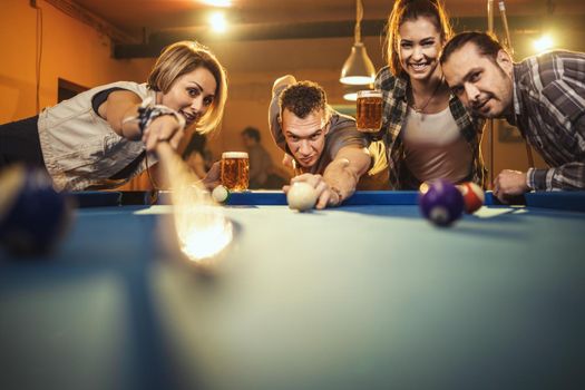 Group of a four smiling cheerful friends are playing billiards in bar after work. They are involved in recreational activity.
