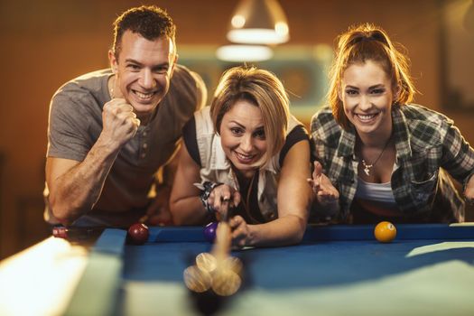 Group of a four smiling cheerful friends are playing billiards in bar after work. They are involved in recreational activity.