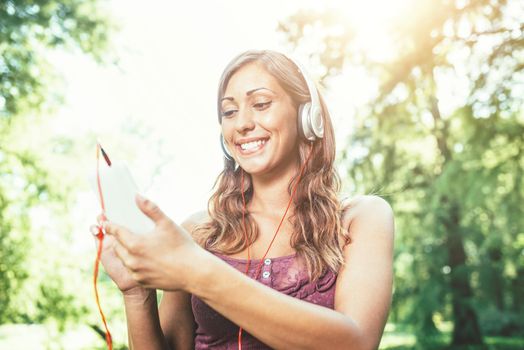 Woman relaxes with headphones listening to music sitting in park. She is doing selfie on her smartphone