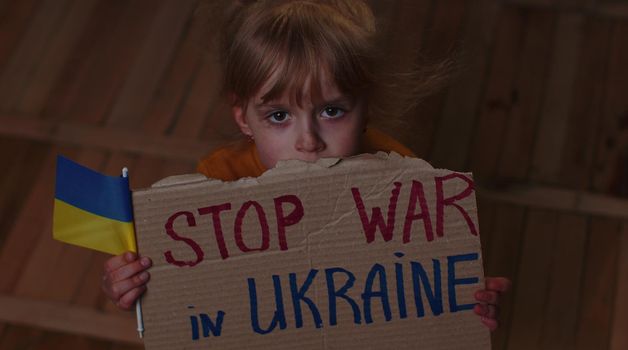 Afraid Ukrainian homeless toddler girl sitting on floor at home holding inscription massage Stop War In Ukraine. Child alone hiding from bombing attack. Crisis, peace, no war, Russian aggression