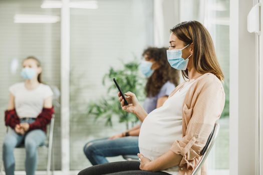 Young pregnant woman with a face mask sitting in a waiting room of a hospital and surfing at her smartphone.