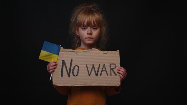 Upset Ukrainian poor toddler girl kid homeless protesting war conflict raises banner with inscription massage text No War on black background. Crisis, peace, stop aggression, child against Russian war