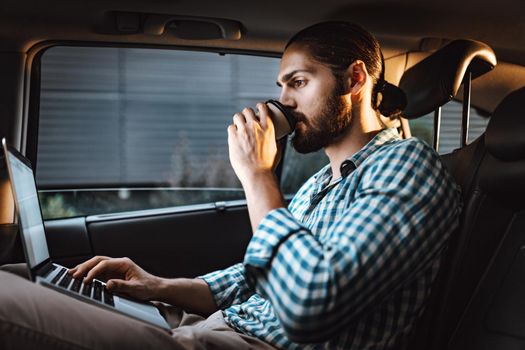 Young confident businessman is holding a cup of coffee in his hand, drinking and texting on a laptop on the back seat in car.
