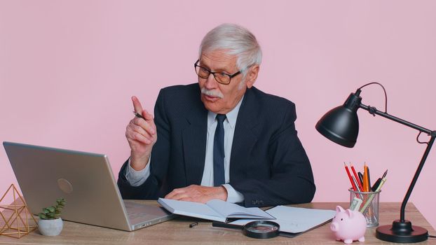 Thoughtful senior businessman making notes, writing down thoughts with pen into notepad notebook diary, to do list, good idea sits at workplace desk at office. Elderly man on pink studio background