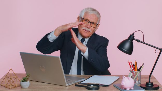 Tired serious upset senior businessman showing time out gesture, limit or stop sign, no pressure, i need more time sits at workplace desk at office. Elderly man isolated on pink studio wall background