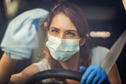 A young woman with a mask on her face  and protective gloves on her nands wipes a car window in her car with a cloth with disinfectant liquid.