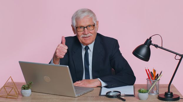 Senior business man employee using laptop computer notebook, raises thumbs up agrees with something or gives positive reply recommends advertisement likes good sits at workplace desk at pink office