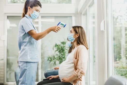 Nurse measuring temperature of a young pregnant woman in waiting room during coronavirus pandemic.