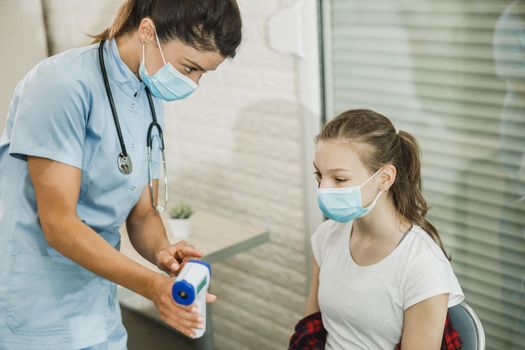 Nurse measuring temperature of a cute female teenager in waiting room during coronavirus pandemic.