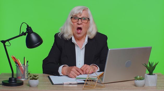 Upset senior businesswoman making face palm gesture, feeling bored, disappointed in result, bad news sits at workplace desk at office. Elderly mature woman isolated indoor on green chroma key studio