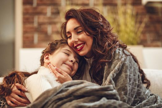 The young mother and her little daughter sit on the sofa and enjoy. Mother is hugging her daughter and smiling, and daughter closed her eyes and cuddle.