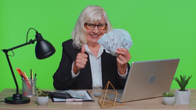 Senior old business woman accountant wears glasses, formal suit, celebrating business success dancing with stack of money dollar cash. Elderly grandmother banker freelancer in green chroma key office