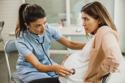 Gynecology nurse talking to young pregnant woman and listening to the patients belly with a stethoscope.