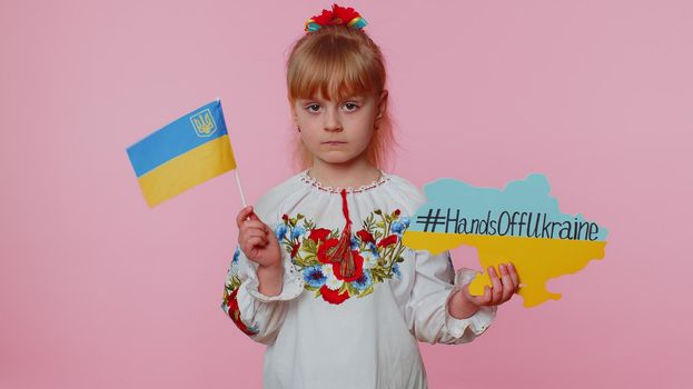 Upset toddler Ukrainian girl kid in embroidery dress protesting war conflict raises banner with inscription massage text on map Hands Off Ukraine on pink background. Peace, no war, stop aggression