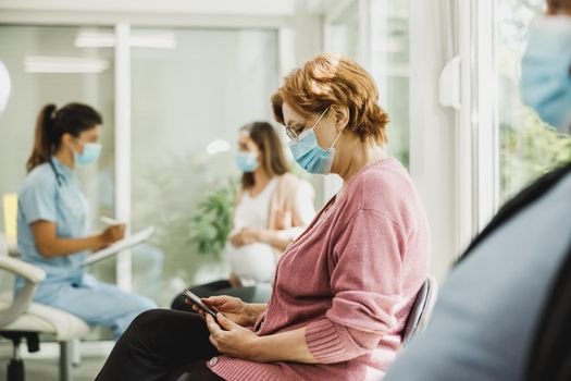Senior woman with a face mask sitting in a waiting room of a hospital and surfing at her smartphone.